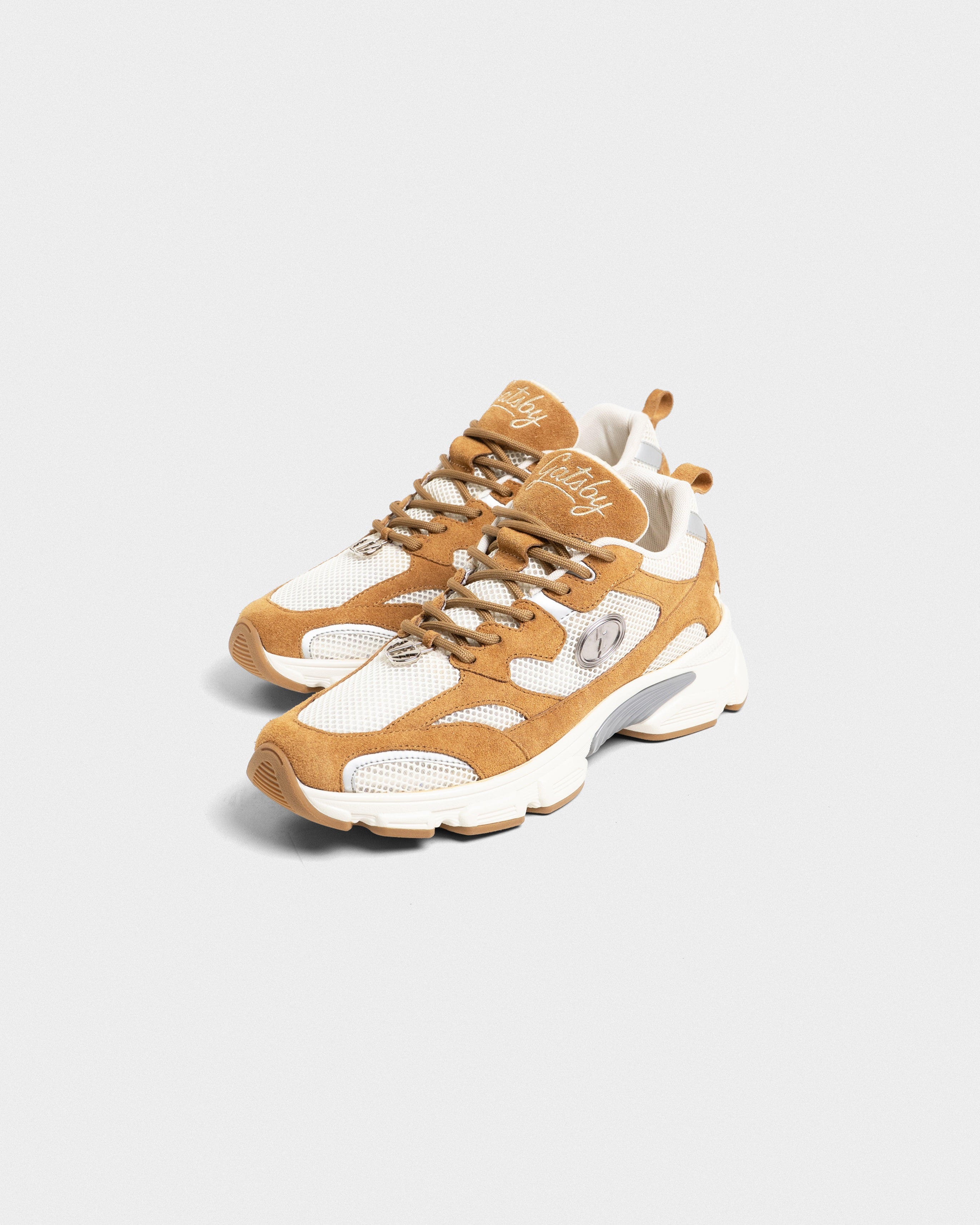 A pair of low-top sneakers in tan and white, featuring mesh panels, suede overlays, chunky laces, and a thick sculpted sole, shown on a light gray background.
